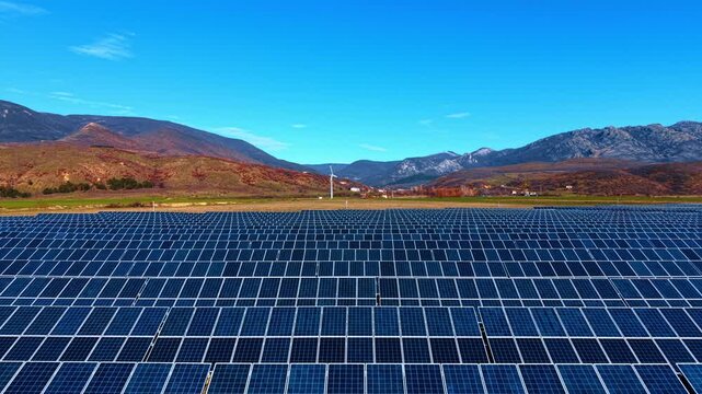Large field of solar panels turned to the sunlight. Wind turbine rotates in the backdrop near the mountains.