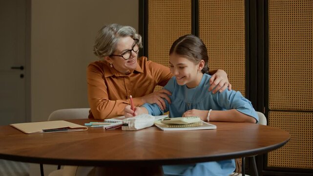 Medium long shot of Caucasian grandmother supporting young girl sitting next to her and doing homework at wooden table in modern apartment