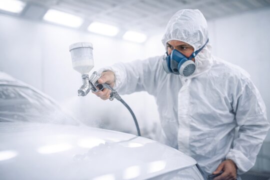 Professional auto painter spraying white car in protective suit inside modern paint booth