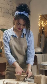 Woman wearing apron shaping clay with hands and smiling. Female working with soft material and forming pottery. Enjoying creative process and focusing on task. Standing in workshop with tools.