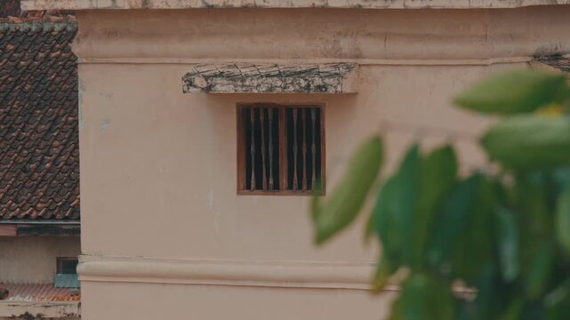 A close-up shot of an ornate weathered window with dark wooden bars set in a pale beige stucco wall of a historic building, captured with a soft cinematic aesthetic.