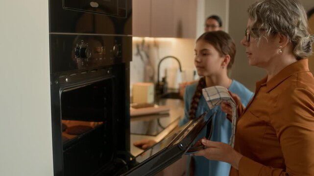 Side view of Caucasian grandmother taking cookies out of oven while baking with granddaughter, mother washing dishes in background in home kitchen
