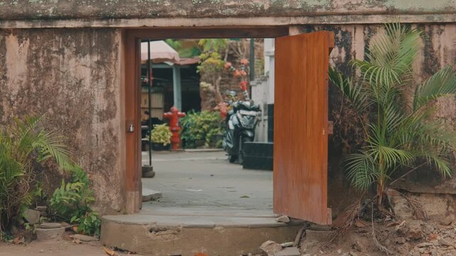 An old weathered wooden door slowly swings open to reveal a hidden Indonesian courtyard with a motorcycle, lush greenery, and a red fire hydrant in the bright daylight.