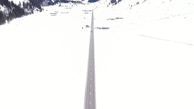 Drone flying above a straight alpine road as a car approaches from the opposite direction, surrounded by bright snowy landscape