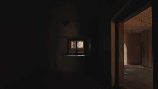 Sunlight streams through a small wooden window into a dark, abandoned room within an old stone structure, highlighting the rough textures and dusty, atmospheric surroundings.
