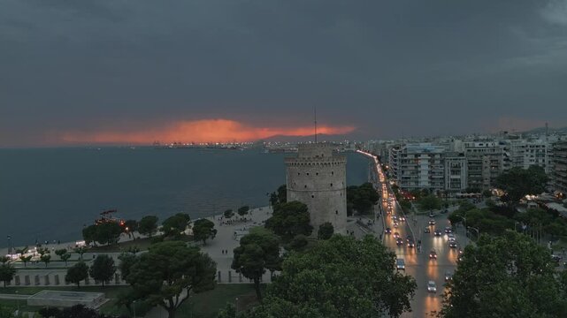 Dynamic aerial hyperlapse over the White Tower of Thessaloniki at dusk. Fast drone flyover showing city traffic and the Thermaic Gulf, Greece.