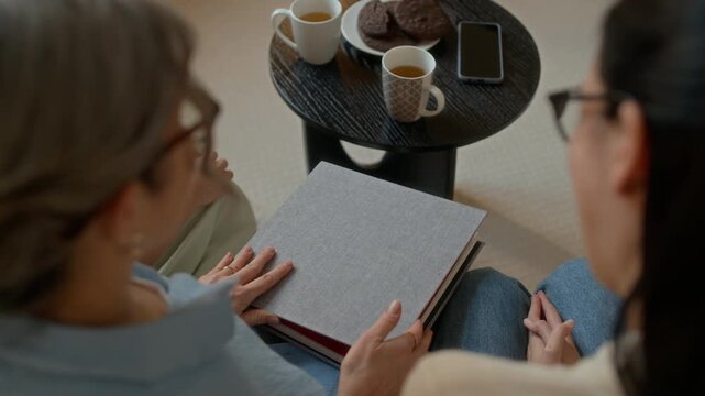 High angle over the shoulder view of woman watching photo album with adult daughter sitting on couch by table with tea and cookies in living room