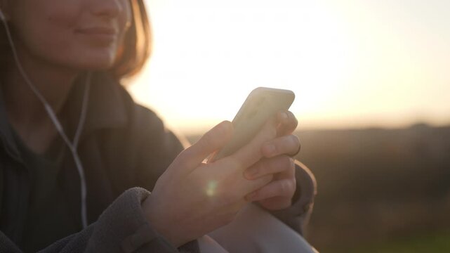 Close up of hands holding smartphone at sunset, woman blurred in background listening to music with wired headphones, wind in hair, warm glowing lens flare, calm everyday moment.