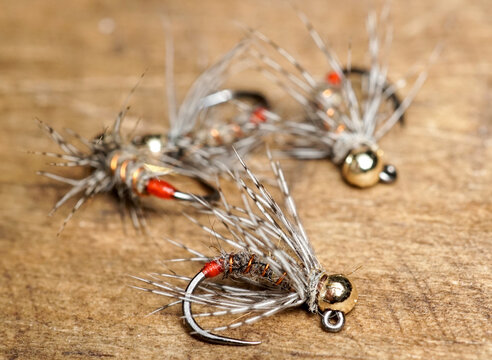 A close-up macro shot of  handmade Fire Orange Tag euro nymph flies with gold tungsten beads and partridge soft hackle, resting on a rustic wooden surface