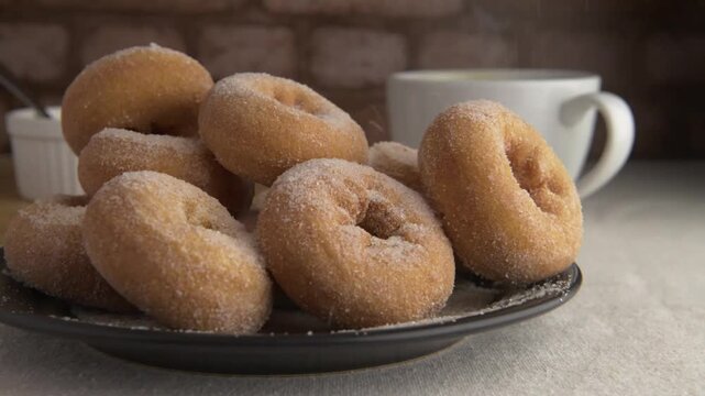 Reposteria casera, donas hechas en casa con azucar cayendo de arriba para cubrirlas de dulce, con una taza de chocolate caliente o cafe humeanate y una pared de ladrillos rustica en el fondo.
