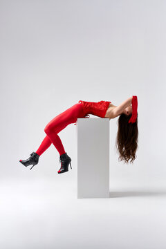 Sexy young woman in red corset and stockings posing on white cube