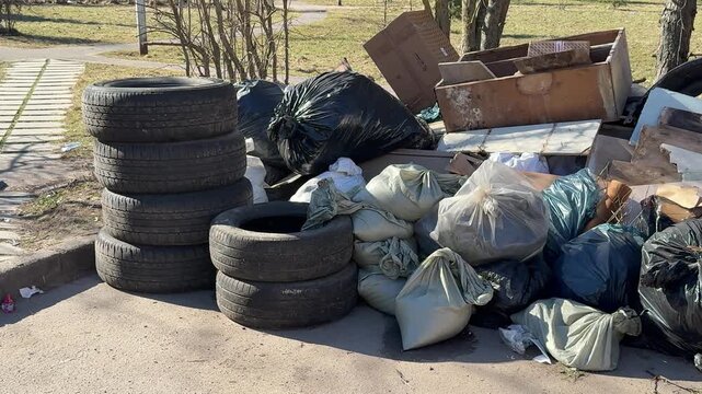 Piled tires and scattered garbage bags on sidewalk, old car tires stacked beside torn bags and cardboard, spring sunlight.
