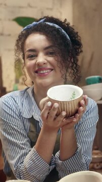 Woman smiling and holding ceramic bowl while showing result. Female presenting handmade pottery and looking at camera. Feeling proud of creation and enjoying work. Working in workshop.