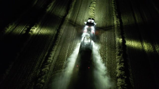 Agricultural baler attached to tractor working at night with illuminated lights captured as aerial panorama during hay or straw processing modern technology