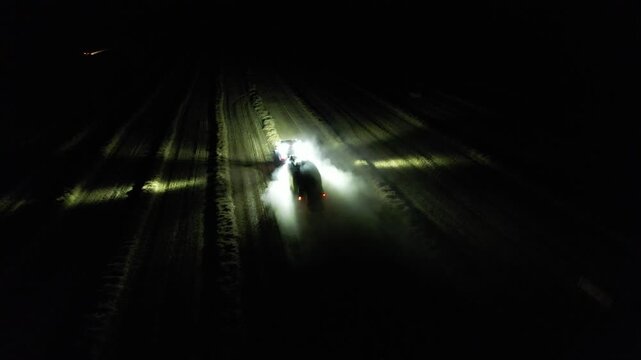Agricultural baler attached to tractor working at night with illuminated lights captured as aerial panorama during hay or straw processing modern technology
