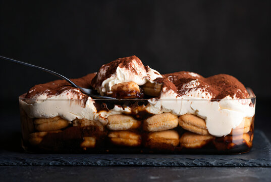A fork is digging into a glass dish filled with tiramisu, showing layers of ladyfingers, cream, and cocoa powder. The dessert is dusted generously with cocoa powder and has a fluffy cream topping.