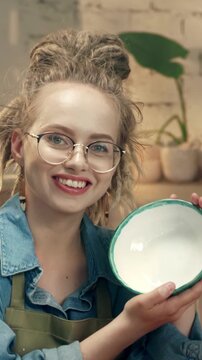 Woman with glasses holding ceramic bowl and smiling at camera. Female presenting handmade pottery and showing result. Feeling proud of creation and enjoying process. Vertical orientation.