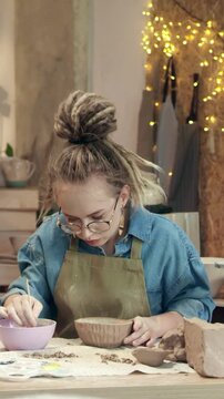 Pretty woman painting ceramic bowl with brush. Female artisan decorating handmade pottery and looking down at work. Creating design. Another person holding finished piece in background.