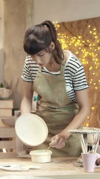 Woman holding ceramic bowl while painting surface with brush. Female artisan working with pottery and looking down at handmade piece. Adding finishing details to crafted object. Creating design.