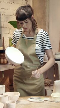 Vertical view of woman holding ceramic bowl while painting inside surface with brush. Female artisan working carefully on pottery and looking down at piece. Creating design details in workshop.