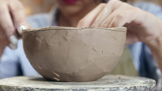 Close view of clay bowl being shaped on wheel. Hands smoothing surface and adjusting edges. Forming ceramic item and refining texture. Working in pottery workshop. Sculpting process.