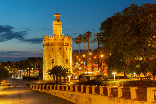 Evening view of Torre del Oro illuminated at dusk with palm trees and city lights in Seville, Spain.