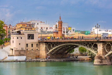 View of Triana Bridge crossing the Guadalquivir River with riverside buildings and city details in Seville, Spain under cloudy sky.