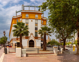 View of a historic yellow and white riverside building with palm trees near Torre del Oro in Seville, Spain under cloudy sky.