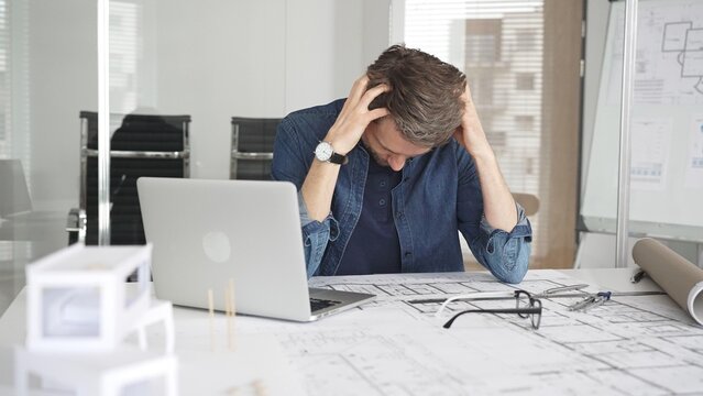 Stressed architect feeling overwhelmed and frustrated, suffering from burnout while working on blueprints for a tight project deadline in a modern office setup