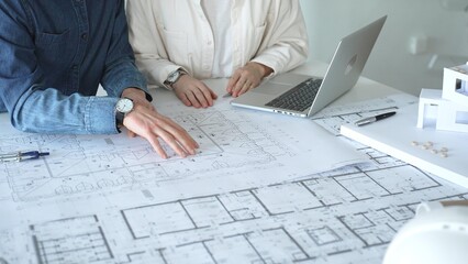 Two architects collaborate over blueprints and a model, using a compass and laptop to measure and plan detailed floor plans, symbolizing teamwork in property development © volha_r