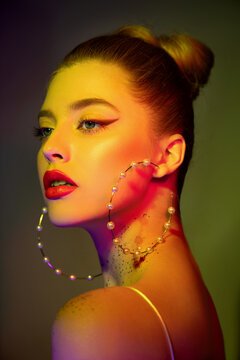 Beautiful young woman  with perfect skin and colourful makeup in neon studio light. Color neon portrait of beauty model with makeup, long eyelashes and big pearl earrings. Selective focus.