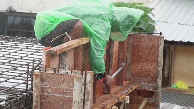 Construction worker in green raincoat working on steel reinforcement during rain. Industrial labor and building site development under difficult weather conditions concept. asian worker