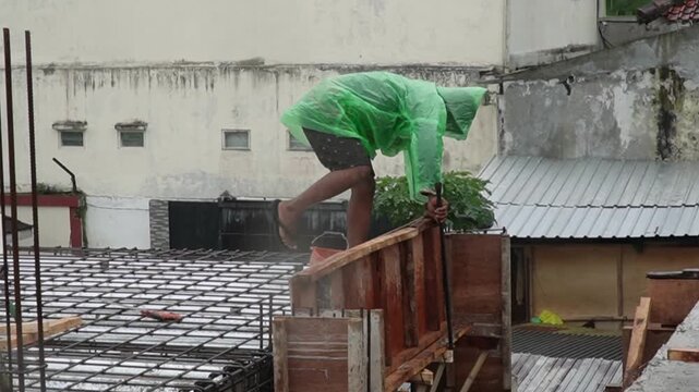 Construction worker in green raincoat working on steel reinforcement during rain. Industrial labor and building site development under difficult weather conditions concept. asian worker