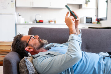 Fototapeta premium Man lying on sofa using smartphone with serious expression, focused and bored. Indoor lifestyle scene showing device dependence, scrolling, notifications and solitary digital consumption at home