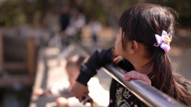 Little girl gazes over railing with curiosity. She holds rail firmly, leaning forward. Sunlight glints off metal as she watches. Her ponytail holds a purple flower clip. She wears dark, patterned top