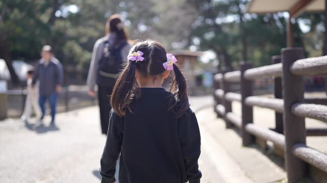 Little girl walks with her mother through a park. Pink hair clips brighten her dark hair. Mother walks slightly ahead, gently guiding her. Sunlight dapples the path behind them