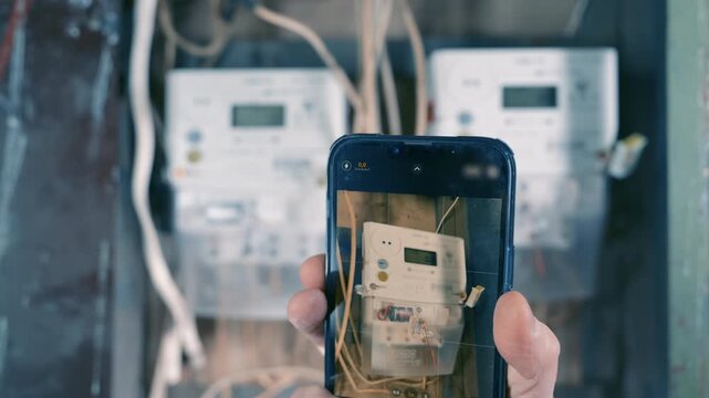 Hand with Smartphone Photographing Two Meters in Large Main Electrical Panel | User Capturing Utility Readings on Phone | Multi-Family Residential Power Consumption Check Concept