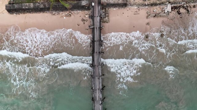 Overhead riser drone shot of a long wooden dock centered over a sandy beach at Liminangcong with waves crashing below, Philippines