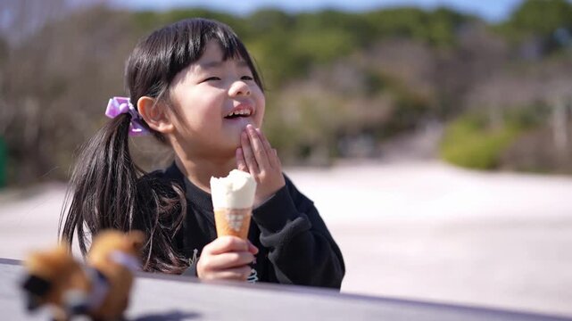 Little girl savors ice cream outdoors. She smiles gently, enjoying sweet treat. Soft sunlight bathes her dark hair and face. Trees and gravel path frame the natural setting