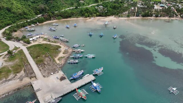 Pull away tilt-up drone shot of San Fernando Ruru Port in El Nido, Palawan, showing expedition outrigger boats in calm turquoise waters, Philippines