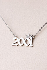 Close-up of a silver pendant with the year 2001 and a cute crown on a chain, set against a luxurious white leather background.