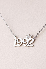 Close-up of a silver pendant with the year 1992 and a cute crown on a chain, set against a luxurious white leather background.