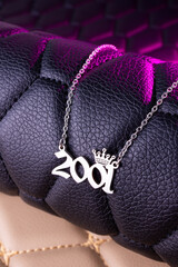 Close-up of a silver pendant with the year 2001 and a cute crown on a chain, set against a luxurious black leather background.
