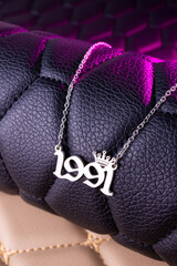 Close-up of a silver pendant with the year 1991 and a cute crown on a chain, set against a luxurious black leather background.