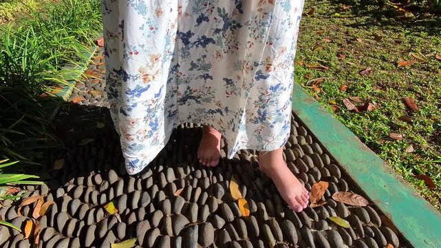 Video of a woman's feet in a long white skirt stepping on a path with small stones to massage reflexology points. 4K footage. Traditional foot therapy with stepping on pebbles.