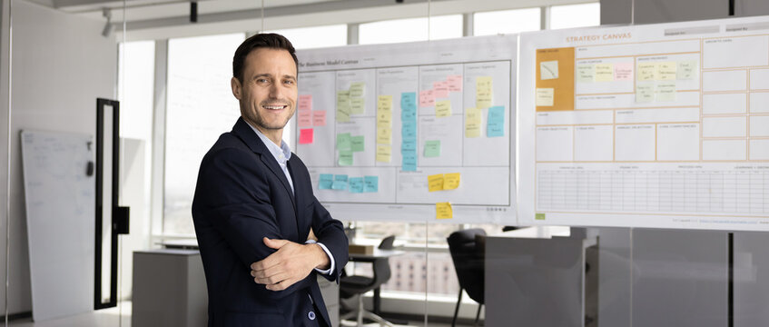 Modern planning culture. Happy confident male business professional project leader agile coach pose in office near sticky note roadmap strategy canvas look at camera. Wide image for horizontal banner