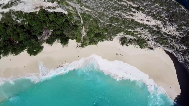 Aerial top-down view of Kelingking Beach, Nusa Penida, Bali. Stunning turquoise ocean waves crash onto a pristine white sand beach bordered by dramatic green cliffs. Perfect for travel and nature