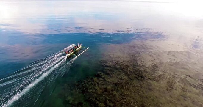 Aerial view of a traditional Balinese outrigger boat gliding over a crystal-clear coral reef. Calm turquoise waters reflect the soft morning sky in a serene tropical setting.