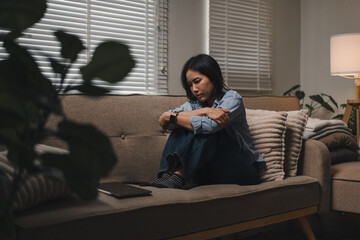 Sad young Asian woman sitting on a sofa hugging her knees in a dimly lit living room © PRIME STOCK LAB