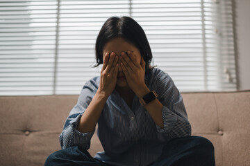 Stressed young woman covering her eyes while sitting on a couch in a dim living room © PRIME STOCK LAB